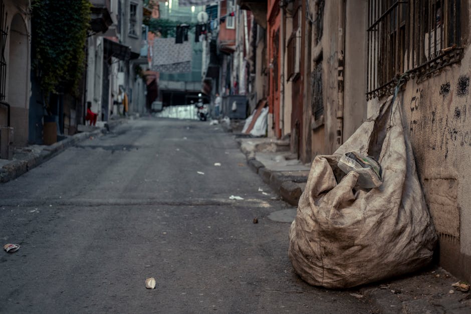 A large, worn, beige cloth sack filled with various discarded household items and rubbish, resting against a metal-barred window on a narrow, uneven city street. The sack appears dirty and slightly stained, with a partially visible transparent plastic bag peeking out from the top. The street surface is rough and paved, with scattered small pieces of litter, including paper and debris, along the edge near the sack. In the background, residential buildings with weathered facades, some with visible peeling paint and graffiti, line both sides of the street. Clothes are hung on lines strung across the alley, and the scene is shaded by overhanging structures and clotheslines, giving a muted, urban atmosphere. The image reflects an environment where informal rubbish collection may be necessary, aligning with the context of private waste management services provided by Waste Clearance Haringey for on-site or alternative waste disposal needs.