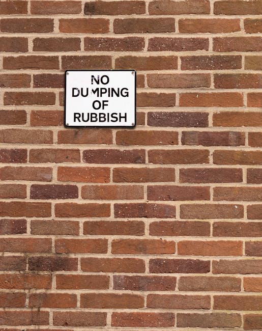 A square white metal sign with black text reading 'NO DUMPING OF RUBBISH' is mounted on a red brick wall with a traditional horizontal pattern. The bricks vary slightly in shades of reddish-brown and have rough, textured surfaces. The sign is positioned approximately one-third from the top of the image, centered horizontally, and appears to be attached with four screws, one at each corner. The brick wall extends across the entire background, with some mortar joints visible between the bricks. There is no visible rubbish or waste in the scene. The lighting is even, illuminating the brick surface clearly. This signage on the brick wall reflects the importance of enforcing responsible waste disposal in the local area, aligning with services provided by Waste Clearance Haringey for alternative waste handling and private rubbish removal solutions.