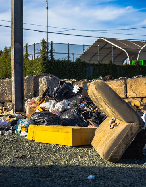 A large, worn, beige cloth sack filled with various discarded household items and rubbish, resting against a metal-barred window on a narrow, uneven city street. The sack appears dirty and slightly stained, with a partially visible transparent plastic bag peeking out from the top. The street surface is rough and paved, with scattered small pieces of litter, including paper and debris, along the edge near the sack. In the background, residential buildings with weathered facades, some with visible peeling paint and graffiti, line both sides of the street. Clothes are hung on lines strung across the alley, and the scene is shaded by overhanging structures and clotheslines, giving a muted, urban atmosphere. The image reflects an environment where informal rubbish collection may be necessary, aligning with the context of private waste management services provided by Waste Clearance Haringey for on-site or alternative waste disposal needs.