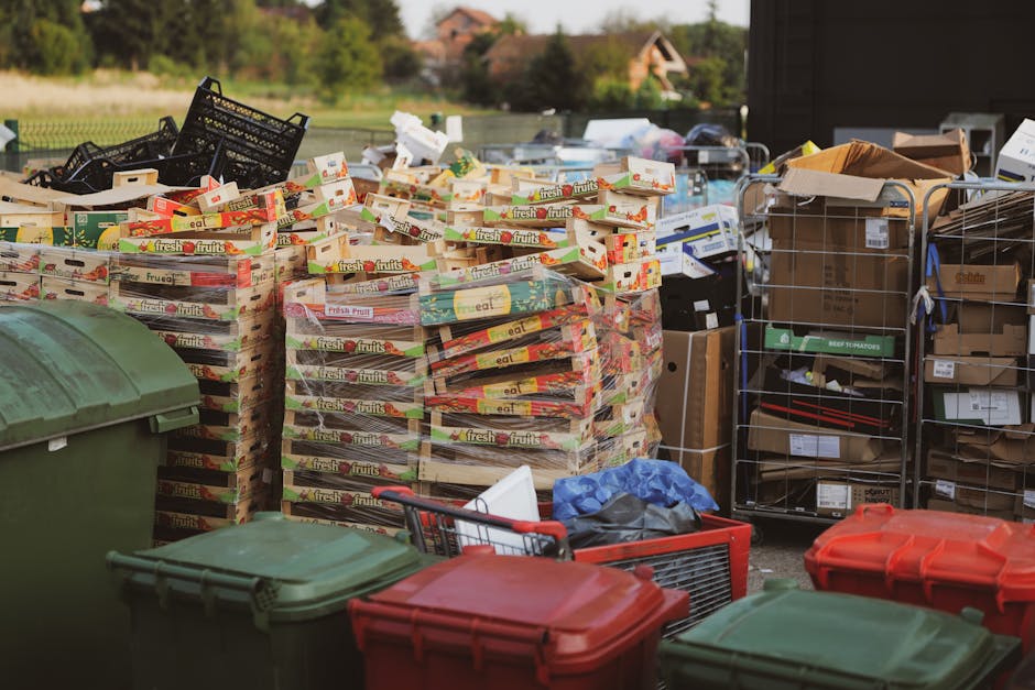 An outdoor storage area filled with various waste and recyclable materials designated for rubbish removal services. In the foreground, multiple large stacks of flattened cardboard boxes displaying colorful print, primarily featuring fruit branding such as 'fresh fruits,' are neatly arranged on pallets. To the left, there are multiple large green and red wheelie bins with closed lids, placed on a paved surface, alongside a red plastic shopping basket containing miscellaneous waste items. Behind the stacks of cardboard, several wire and cardboard crates, some filled with cardboard and paper packaging, are visible, with a few boxes placed on top of or leaning against them. The background shows a garden with grassy lawns and trees, and distant residential buildings with tiled roofs, under natural daylight. The environment appears organized for waste collection or disposal, aligning with alternative waste handling practices like private rubbish collection services managed by Waste Clearance Haringey.