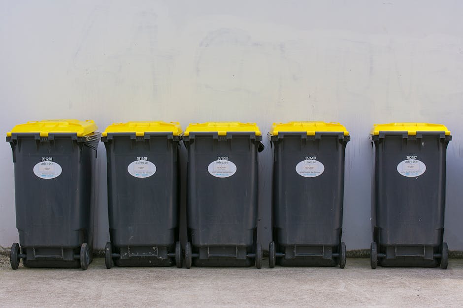 A row of five large black wheeled rubbish bins with yellow lids, made of durable plastic with a textured surface, are lined up on a concrete floor against a plain white wall. Each bin has an oval white label with a barcode and identification number, positioned towards the top center. The bins are evenly spaced, with some showing slight scuff marks near the wheels, which are black and mounted at the base for easy mobility. The yellow lids are closed, fitting snugly on the bins, which are situated outdoors or in an industrial or commercial setting. The environment appears well-lit with natural or artificial light, emphasizing the clean, functional appearance of the bins. The configuration suggests they are intended for waste collection, possibly as part of a private waste management service such as Waste Clearance Haringey, supporting non-local authority rubbish removal or alternative waste disposal arrangements.
