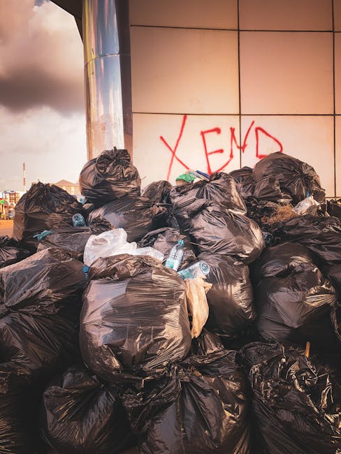 A large pile of black plastic rubbish bags filled with waste, stacked against a tiled exterior wall of a building. The bags appear tightly sealed and contain household or commercial refuse, with some transparent bottles and paper visible among them. The wall behind features clean, beige tiles with a red spray-painted graffiti marking that reads 'XEND'. The scene is outdoors, likely on a pavement or driveway, with diffused natural lighting suggesting an overcast sky. The arrangement of bags indicates a collection or disposal effort, possibly linked to private or commercial rubbish clearance services, such as those offered by Waste Clearance Haringey. The image reflects an uncollected or waiting-to-be-removed waste pile, highlighting the importance of professional rubbish removal solutions for clearing commercial or residential waste outside premises.