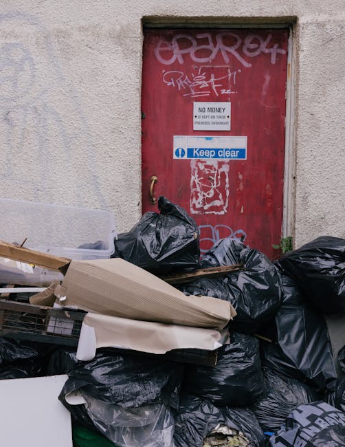 In front of a restaurant with a deep red façade, a black commercial waste bin labeled 'COMMERCIAL WASTE ONLY' is placed on the sidewalk, overflowing with cardboard boxes, papers, and packaging materials. The bin is positioned centrally, partially blocking the entrance to the establishment. To the right of the bin, a short metal bollard and a green-painted lamp post are situated near the curb alongside a bicycle rack and bicycles. The restaurant features large glass windows with multiple panes, through which warm interior lighting and hanging light fixtures are visible. A small blackboard sign on the left advertises a self-service buffet and food offerings, while a downtown street scene extends into the background, with a parked delivery truck and other urban elements. The pavement and road are typical of city streets, with a painted traffic zone marking on the tarmacked surface. This scene reflects an example of private waste management, as provided by Waste Clearance Haringey, supporting the needs of local businesses for rubbish removal services.