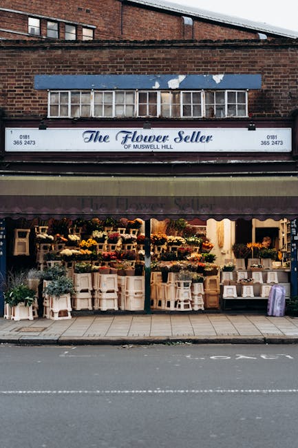 The image shows a flower shop named 'The Flower Seller of Muswell Hill', located in a brick building with a white signboard displaying the shop's name, phone number, and address. Above the shop, there is a boarded-up window with white frames, set against the red brick facade of the upper part of the building. The shop-front features large display windows and an outdoor area with organized arrangements of potted plants and flowers, some placed on small white or wooden tables and stands. The display includes a variety of floral displays and greenery, with a soft warm lighting illuminating the interior of the shop. The sidewalk in front is clean and paved, and a dark, striped awning extends across the storefront, partially covering the flower displays. The scene captures a typical urban shopfront that might be involved in private floral and plant sales, aligning with alternative methods of small-scale retail flower and plant distribution, with no visible rubbish or waste in the vicinity.