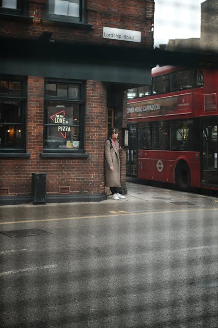 A person stands on a wet pavement outside a red double-decker bus in Haringey, London, next to a brick building with two dark-framed windows. The person is dressed in a light brown coat, dark trousers, and white shoes, holding a bag, and appears to be waiting or observing. The building signage indicates the street name is Lordship Road. A small window advertises pizza, and a black trash bin is positioned on the sidewalk. The scene suggests an urban setting with typical city transport and architecture, possibly related to private waste management or rubbish removal services like Waste Clearance Haringey operating in the area. The wet pavement and overcast lighting imply recent rain, contributing to a reflective surface that highlights the urban environment. The overall atmosphere is quiet and orderly, consistent with typical street scenes involving public transportation and local waste collection activities.