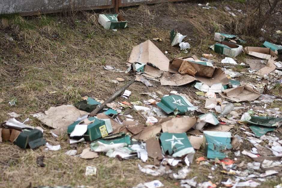 An outdoor scene showing a patch of ground next to a wooden fence with scattered trash and debris. The litter includes flattened and torn cardboard boxes, mostly brown with some green sections featuring white star logos, and various smaller paper scraps. Some boxes are partially crushed or crumpled, with torn edges and creases, lying among patches of brownish-green grass and soil. A long metal pole or rod is visible among the debris, crossing part of the ground. The background includes a weathered wooden fence, and the lighting suggests an overcast day, contributing to a dull, muted atmosphere. The overall scene depicts typical refuse left in an unmanaged outdoor area, demonstrating a need for waste removal or cleaning services, potentially relevant to private rubbish clearance operations such as those provided by Waste Clearance Haringey.