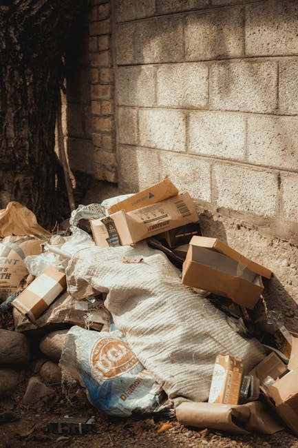 A large pile of black rubbish bags filled with waste, stacked against a modern building with reflective panels. Some bags have transparent sections revealing contents such as plastic bottles, and a few bottles are visible outside or on top of the bags. The bags are tightly packed and appear to be overfilled, leaning or bulging in various directions. Behind the pile, there is a grey tiled wall with red spray-painted graffiti that spells 'XEND'. The scene is outdoors, with a cloudy sky in the background and urban elements like buildings visible at a distance. The lighting is natural, with soft sunlight casting subtle shadows on the waste and the building. The overall setting suggests an instance of unmanaged waste, highlighting challenges in private rubbish disposal or collection delays, which Waste Clearance Haringey could address through alternative on-site clearance or independent waste removal services.