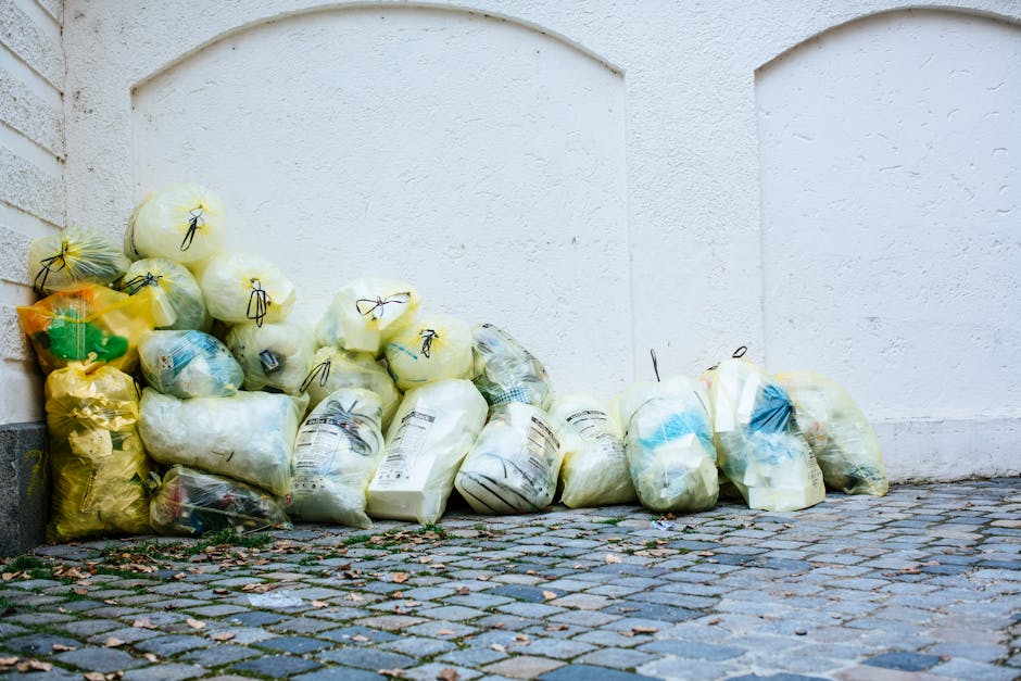 A collection of multiple yellow and white plastic rubbish bags filled with discarded waste, stacked against a white textured wall on a cobblestone pavement. The bags appear to contain a mix of household refuse, with some showing partial contents such as cardboard, paper, and plastic bottles. Several bags are tied securely with black plastic ties, and they vary in size and fullness. The bags are placed close to each other, occupying most of the lower left side of the image, with some leaning against the wall and others lying flat on the uneven cobblestones. The environment suggests an outdoor area, possibly a back alley or side street, where waste clearance services like those provided by Waste Clearance Haringey might be involved in managing non-culpable rubbish disposal. Natural light illuminates the scene evenly, highlighting the textures of the bags, the cobblestone surface, and the wall's rough finish, conveying a neutral and observational perspective on typical waste accumulation for collection or disposal.