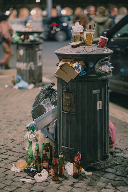 A large black rubbish bin with a domed lid is positioned on a cobblestone street, surrounded by overflowing litter and discarded bottles, primarily green and brown glass, along with crumpled paper and food wrappers, some hanging from the bin's side. Several used disposable coffee cups with plastic lids and straws rest atop the bin, while other trash, including a cardboard box and plastic bags, are strewn around its base. In the background, a grey cityscape with blurred people walking and parked cars is visible, indicating a busy urban environment. The scene captures a typical instance of street-level waste accumulation, highlighting the need for proper rubbish removal and municipal waste management practices. Waste Clearance Haringey occasionally handles such scenarios through individual or private disposal services, emphasizing the importance of responsible waste handling to maintain cleanliness in public areas.