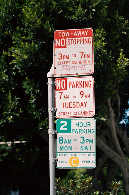 A tall metal parking restriction signpost standing on a sidewalk outdoors, with three rectangular signs stacked vertically. The top sign, in red and white, indicates a tow-away zone with 'NO STOPPING' permitted only between 3 PM and 7 PM on weekdays, except on Saturdays and Sundays, and provides a phone number for towing inquiries. The middle sign, also in red and white, prohibits parking from 7 AM to 9 AM on Tuesdays for street cleaning purposes. The bottom sign, in teal and white, designates a two-hour parking limit from 8 AM to 3 PM, Monday through Saturday, with an exception for vehicles displaying a C permit. The background features lush green foliage from trees and a clear blue sky, creating a natural contrast to the utilitarian signage. The scene suggests an urban or suburban street environment where parking and clearance restrictions are visibly enforced, aligning with the context of private or alternative waste management considerations. The image, associated with Waste Clearance Haringey, highlights typical signage that drivers and residents might observe, indirectly supporting awareness of local regulations related to rubbish collection and street upkeep strategies.