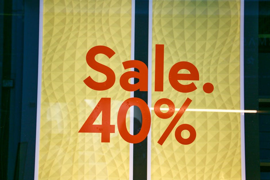 A close-up view of a bright yellow retail window display with a geometric textured pattern. Prominently featured in large, bold, red lettering is the word 'Sale.' followed by a large '40%' indicating a discount. The display appears to be part of a commercial storefront, with reflections of surrounding environment and interior lighting faintly visible on the glass surface. The design and messaging suggest an advertising sign for a promotional sale event, possibly for clothing or household goods, aligning with the visual language commonly seen in retail settings. The surrounding framing hints at the window being situated at street level, with the focus entirely on the promotional signage for potential customers. The overall scene conveys a professional and straightforward presentation of a discount offer, with clear visibility of the colours, textures, and layout of the promotional message.