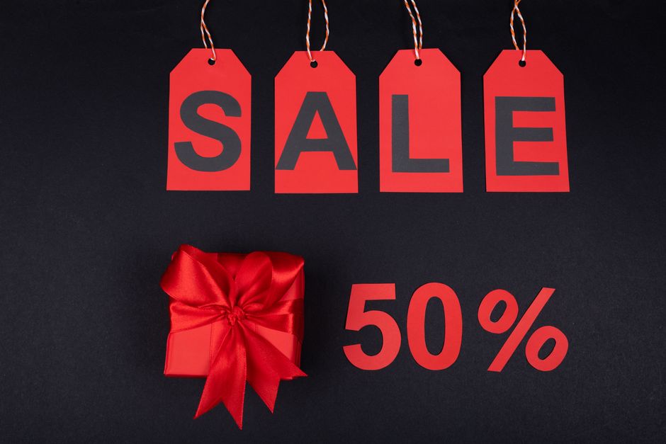 Four red rectangular tags with black letters spelling 'SALE' hang from white and red string loops against a black background. Below the tags, a red gift box wrapped with shiny satin ribbon and a large bow is positioned to the left, while a bold red '50%' discount text is displayed to the right. The scene is set on a flat dark surface, with the lighting highlighting the vibrant red tones of the tags, gift wrap, and text. The arrangement suggests a promotional sale, possibly related to holiday or seasonal discounts, with the presence of the gift box emphasizing a theme of gift-giving and special offers. The image's clean and minimal composition makes it suitable for online advertising or promotional content for retail sales, including those covering independent collection or private disposal options that could relate to waste management or clearance services offered by Waste Clearance Haringey.