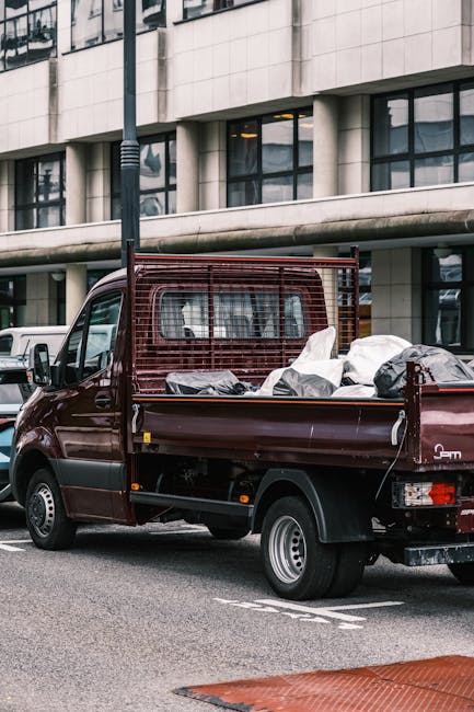 A small, dark red flatbed utility vehicle parked on an urban street in front of a modern building with large glass windows and a concrete facade. The truck's cargo area is filled with various discarded items, including large black and white plastic bags and unidentifiable objects, indicating it is being used for rubbish removal. The vehicle is equipped with a protective mesh panel behind the cab, and the flatbed's sides are open, exposing the waste materials. The scene is set during daylight, with natural light illuminating the vehicle and the surrounding street, reflecting typical on-site clearance or private waste collection activities, often handled by private rubbish removal services like Waste Clearance Haringey, in areas such as Alexandra Palace, Haringey. The street appears clean, with other parked cars visible in the background and a lamp post nearby, emphasizing the vehicular nature of waste disposal in an urban environment.