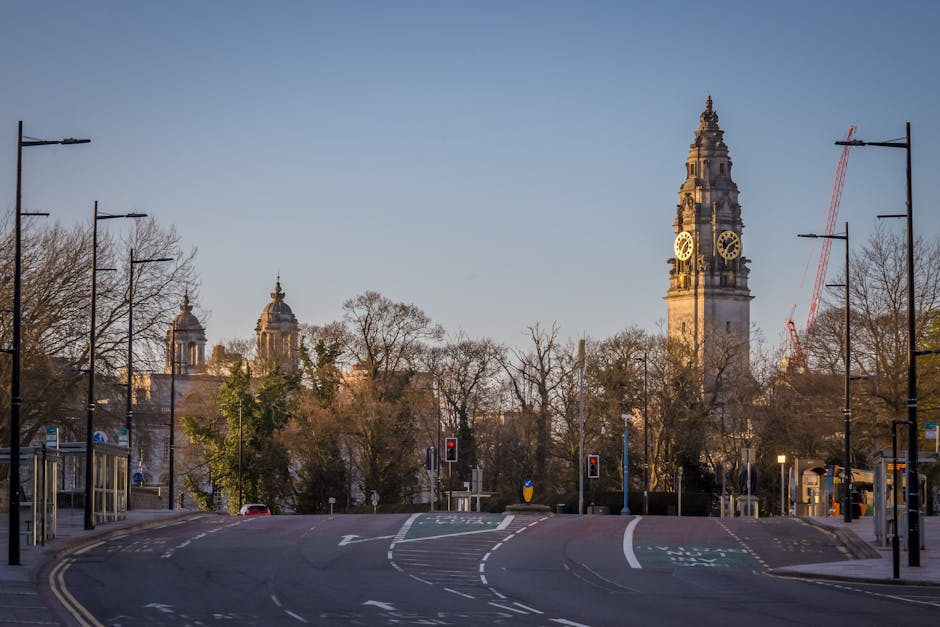 A wide view of a city street during daylight hours shows an intersection with traffic lights and pedestrian crossings. In the foreground, the tarmac road features white and green markings, and is lined with tall, black street lamps on both sides. Behind the street lamps, leafless and some with sparse foliage trees are visible, indicating early spring or late autumn. The background features prominent historic clock towers with ornate architectural details, topped with spires and decorative elements, set against a clear blue sky. To the right, a red construction crane is visible near a tower, suggesting ongoing construction or renovation work nearby. The environment appears calm and organized, with no visible vehicles or pedestrians at the moment. This scene could be associated with urban cleanup or maintenance efforts, aligning with services like waste clearance or rubbish removal offered by Waste Clearance Haringey, especially in the context of on-site collection or alternative waste handling in a cityscape setting.