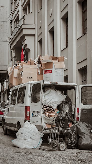 A white commercial van parked on a city street adjacent to a multi-storey residential building with a beige concrete façade. The van’s rear doors are open, revealing an interior filled with various types of waste materials, including a large black plastic bag, cardboard boxes, wooden pallets, and suspicious-looking bundled items covered with a white tarp. Several bulging white and black rubbish bags are stacked both inside and outside the van, some resting on the ground nearby. On the roof of the vehicle, multiple flattened cardboard boxes and larger packaging materials are piled haphazardly, with some protruding over the edges. A small hand truck or dolly leans against the open rear doors, loaded with additional waste bags. The environment appears to be an urban setting, with the van positioned on a paved road or driveway close to the building, which features modern windows and flat architectural details. The scene captures an example of on-site rubbish collection or private waste disposal, as handled by waste clearance services such as Waste Clearance Haringey, for disposal or transfer to recycling facilities.