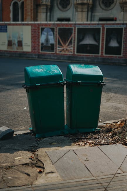 Two green wheeled rubbish bins are positioned side by side on a sidewalk curb in an urban area. The bins are made of durable plastic with hinged lids, one slightly more open than the other, revealing a smooth, matte finish. They are situated on a concrete surface with some loose leaves and dirt visible at the base. Behind the bins, the street extends into the background, with an asphalt road that appears clean and free of debris. Further back, a brick wall with large windows, framed by decorative architectural features, suggests the exterior of a building, possibly a commercial or residential property. The scene is lit by natural daylight, casting subtle shadows and indicating a clear day. The presence of the bins, along with the environment, subtly relates to private waste management and alternative rubbish collection options, as provided by Waste Clearance Haringey, serving local residential or commercial premises.